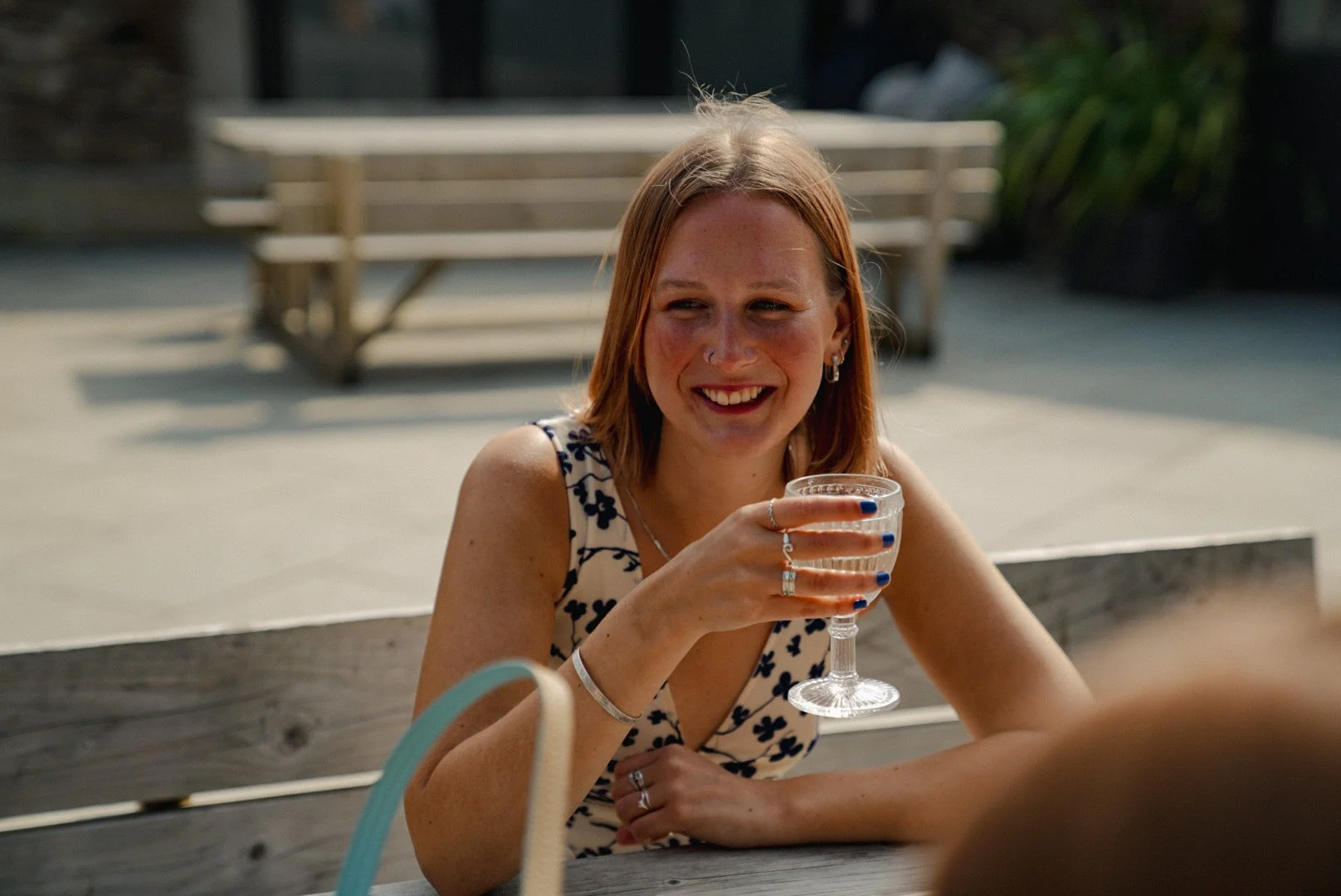A young woman with red hair and a nose piercing, wearing a sleeveless dress with a floral pattern, is smiling and holding a glass of water or cocktail, sitting at a wooden table outdoors.