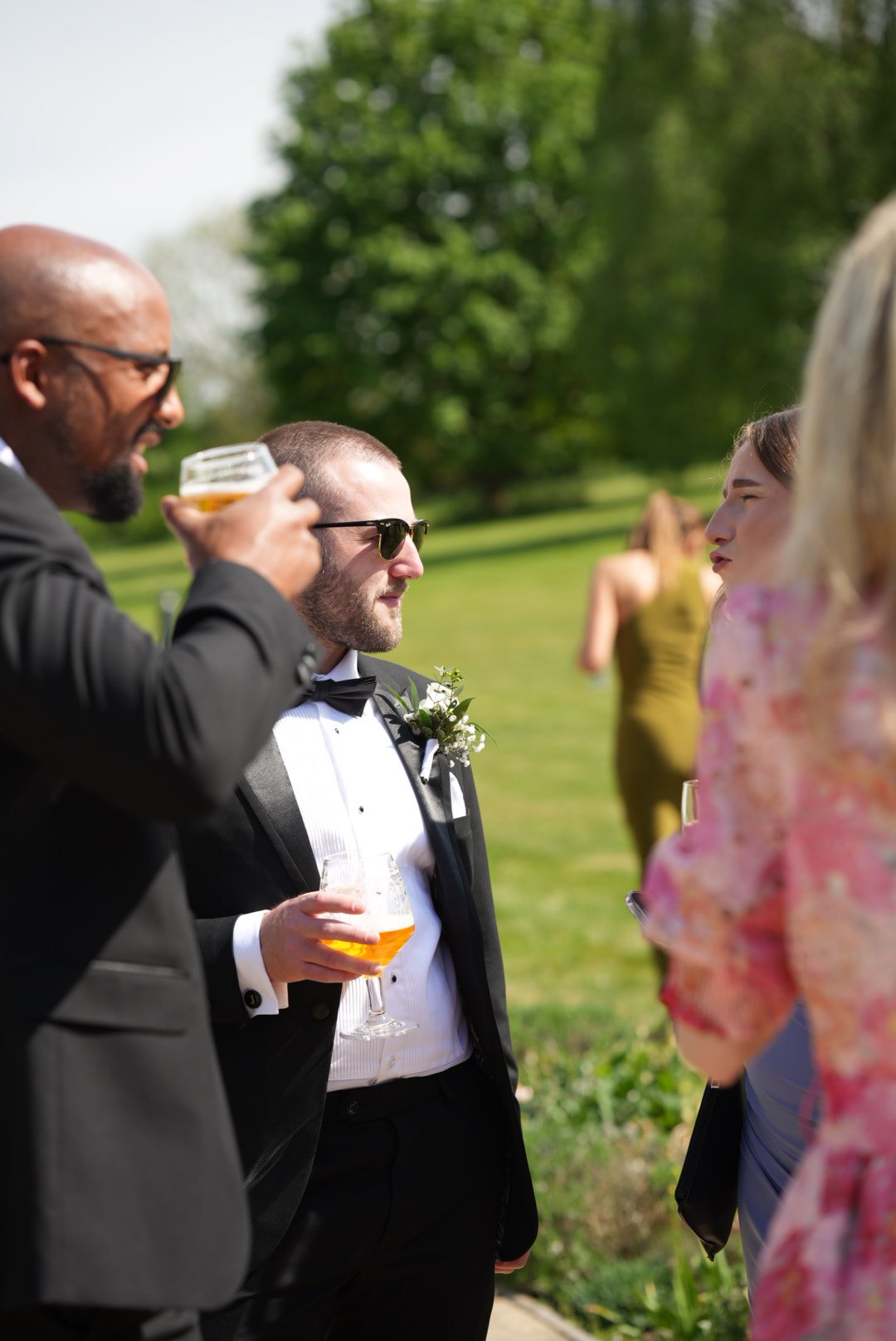 People socializing outdoors on a sunny day, including a man in a tuxedo with a boutonniere holding a glass of beer, and others in the background.