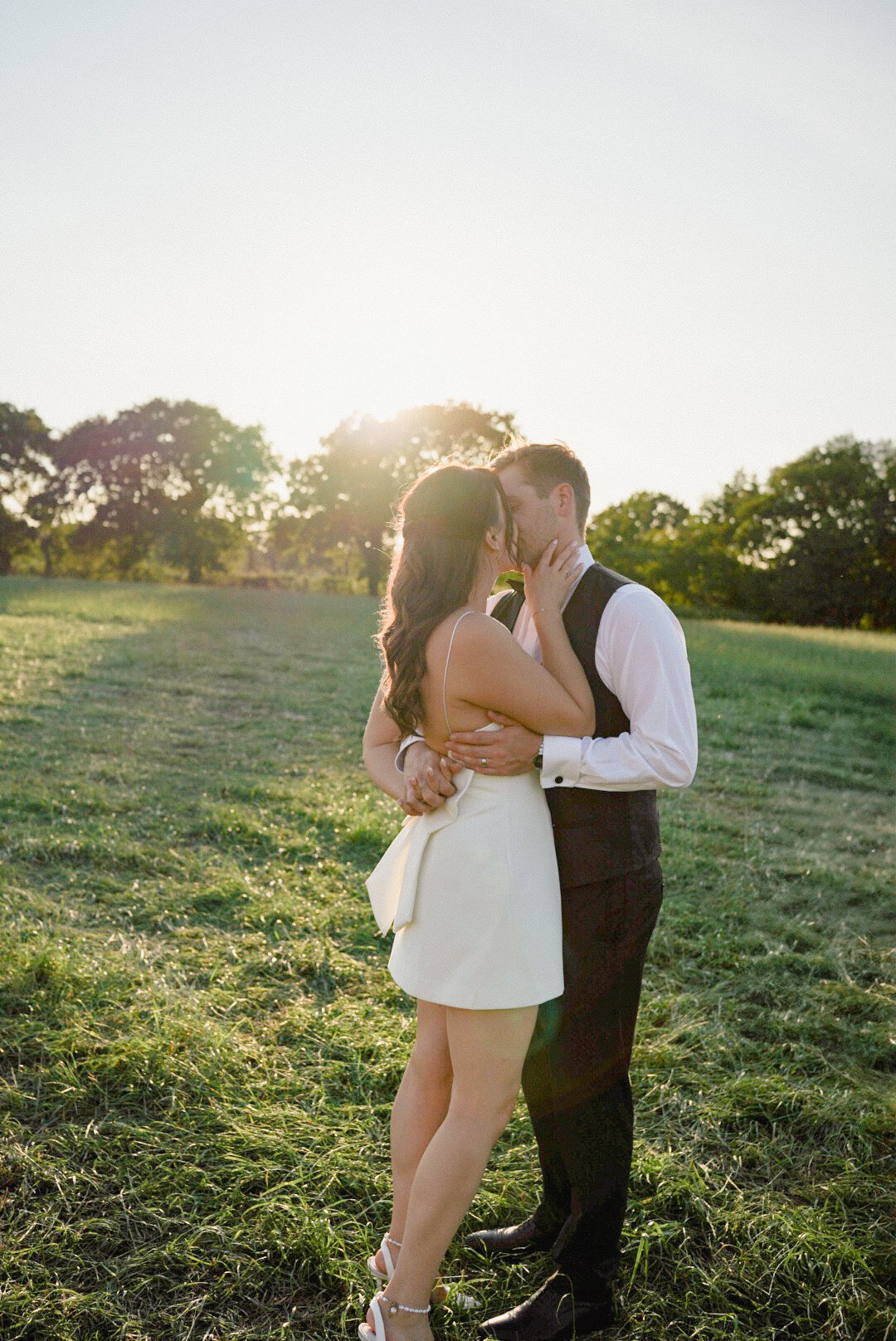A couple kissing in a grassy field with the setting sun behind trees.