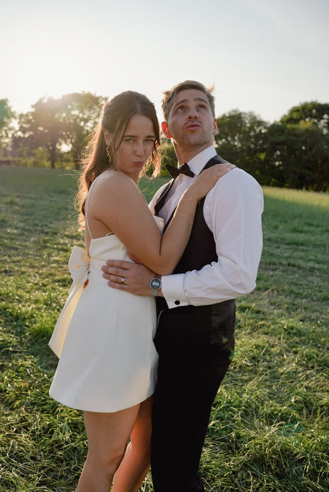A young woman and man, dressed in formal wedding attire, are posing outdoors in a grassy field during sunset. The woman is wearing a white dress with spaghetti straps and has her arm around the man's shoulders. The man is wearing a black suit vest, w