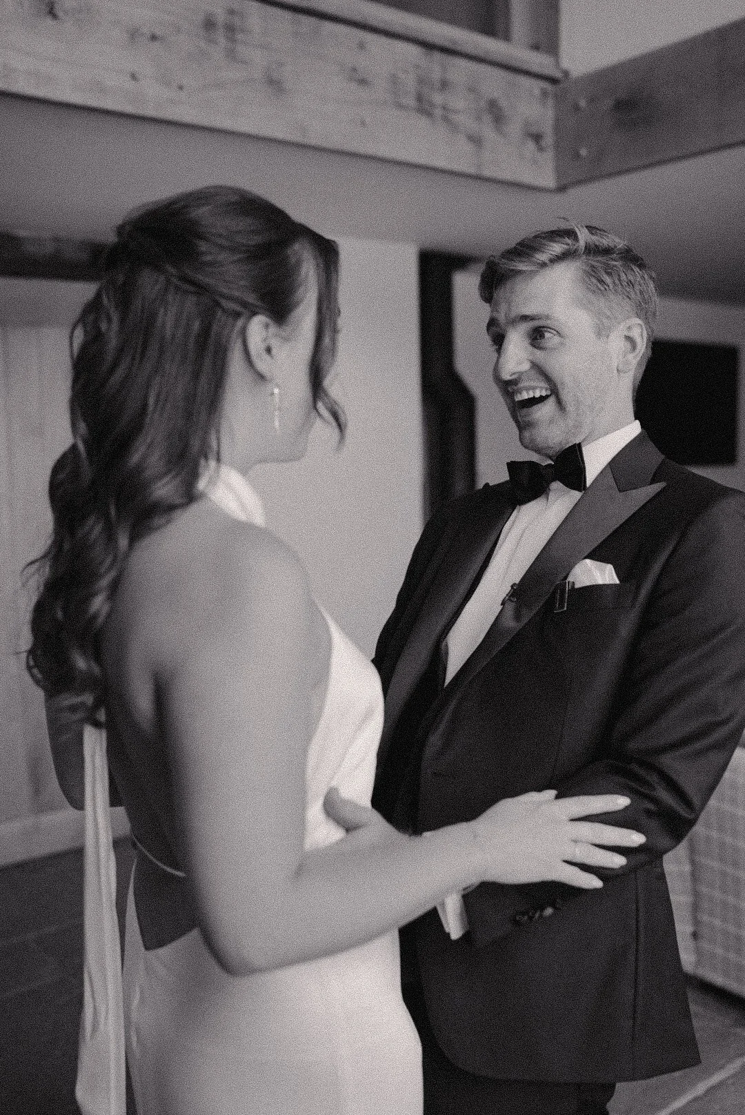A black-and-white photo of a bride and groom smiling and holding each other during their wedding. The groom is wearing a tuxedo with a bow tie, and the bride is in a sleeveless wedding dress with long wavy hair.