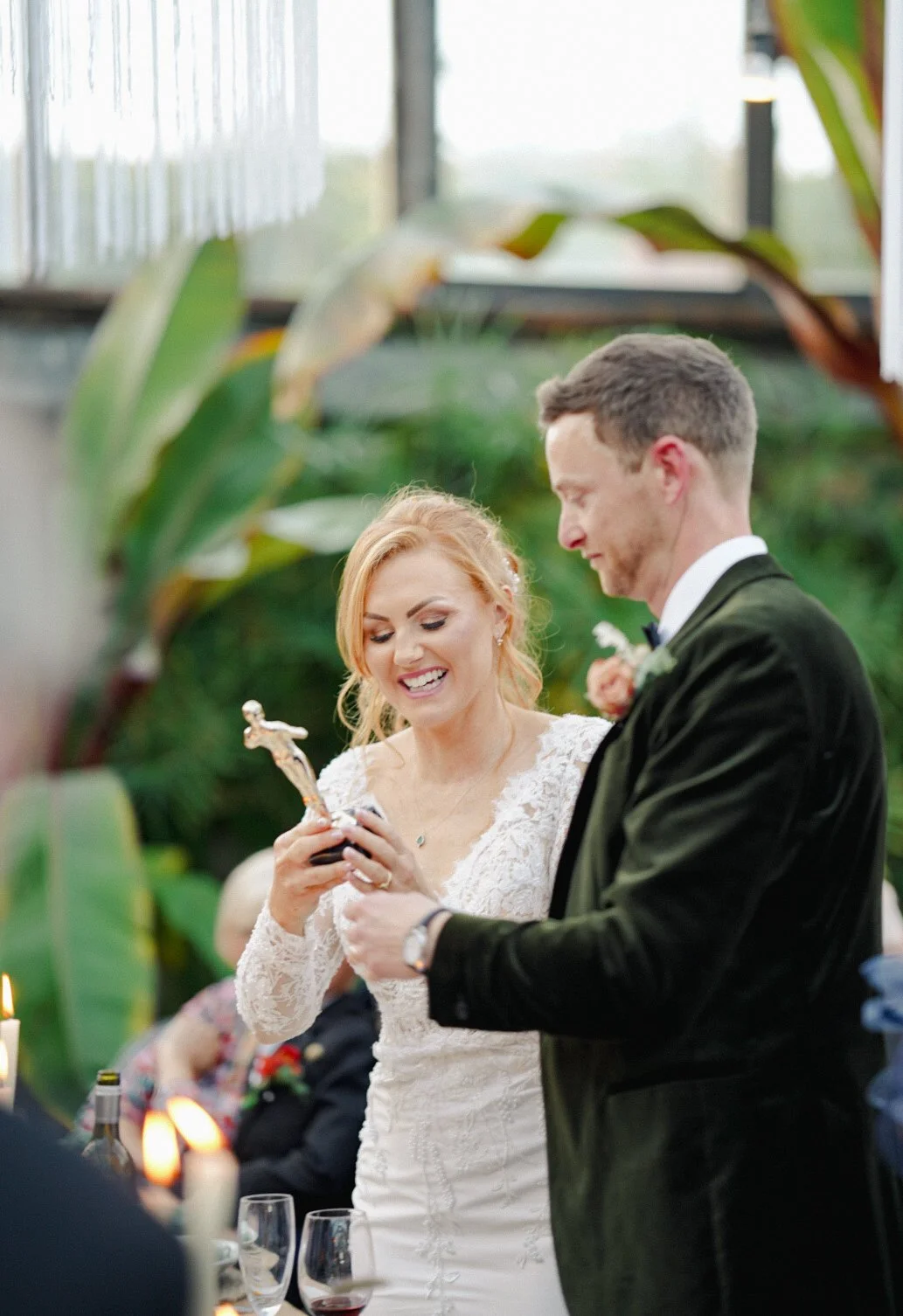 A bride and groom exchanging rings during their wedding ceremony, with the bride holding a small crucifix statue, indoors with greenery in the background.