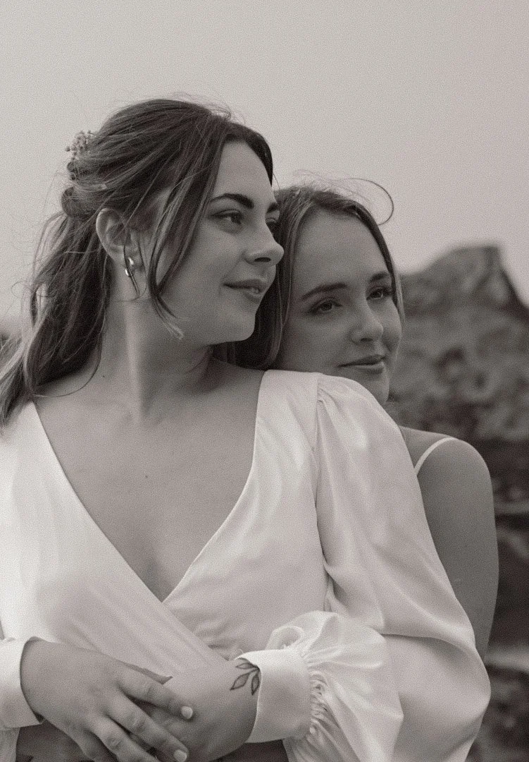 Two young women standing close together outdoors, with a rocky landscape in the background, in black and white.