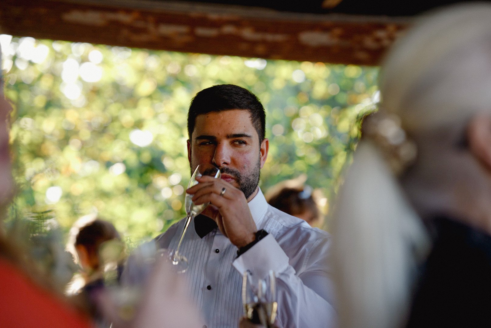 A man in a tuxedo with a striped shirt and black bow tie is holding a champagne flute near his lips, appearing to be taking a sip, at an outdoor event with a blurred background of greenery and other guests.