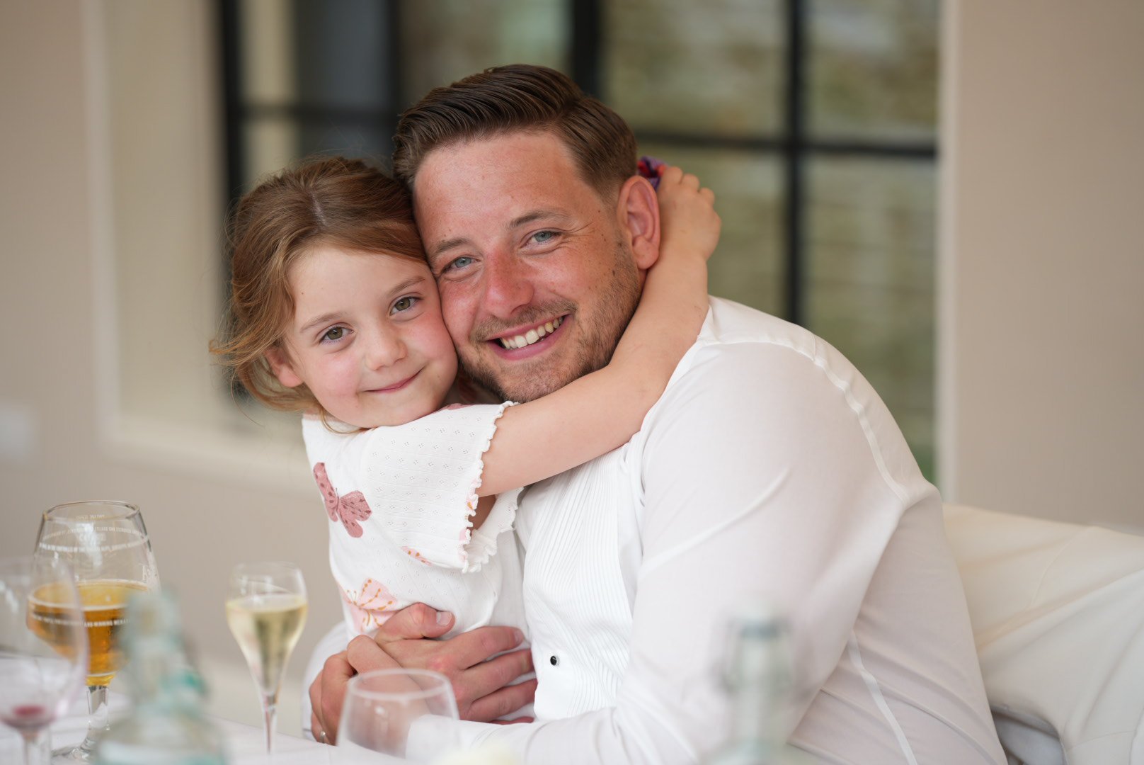 A young girl hugging an adult man, both smiling happily, at a dining table with glasses of wine and drinks in front of them.