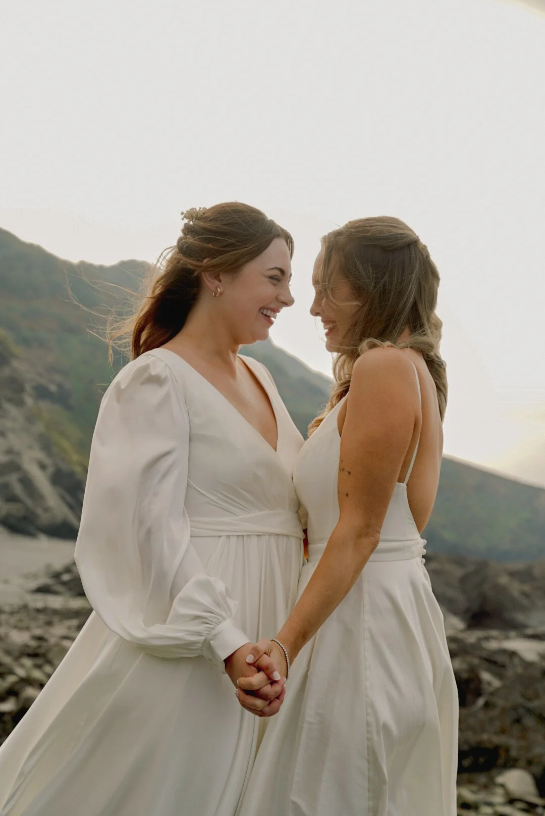 Two women in white dresses holding hands and smiling at each other outdoors with mountains in the background.