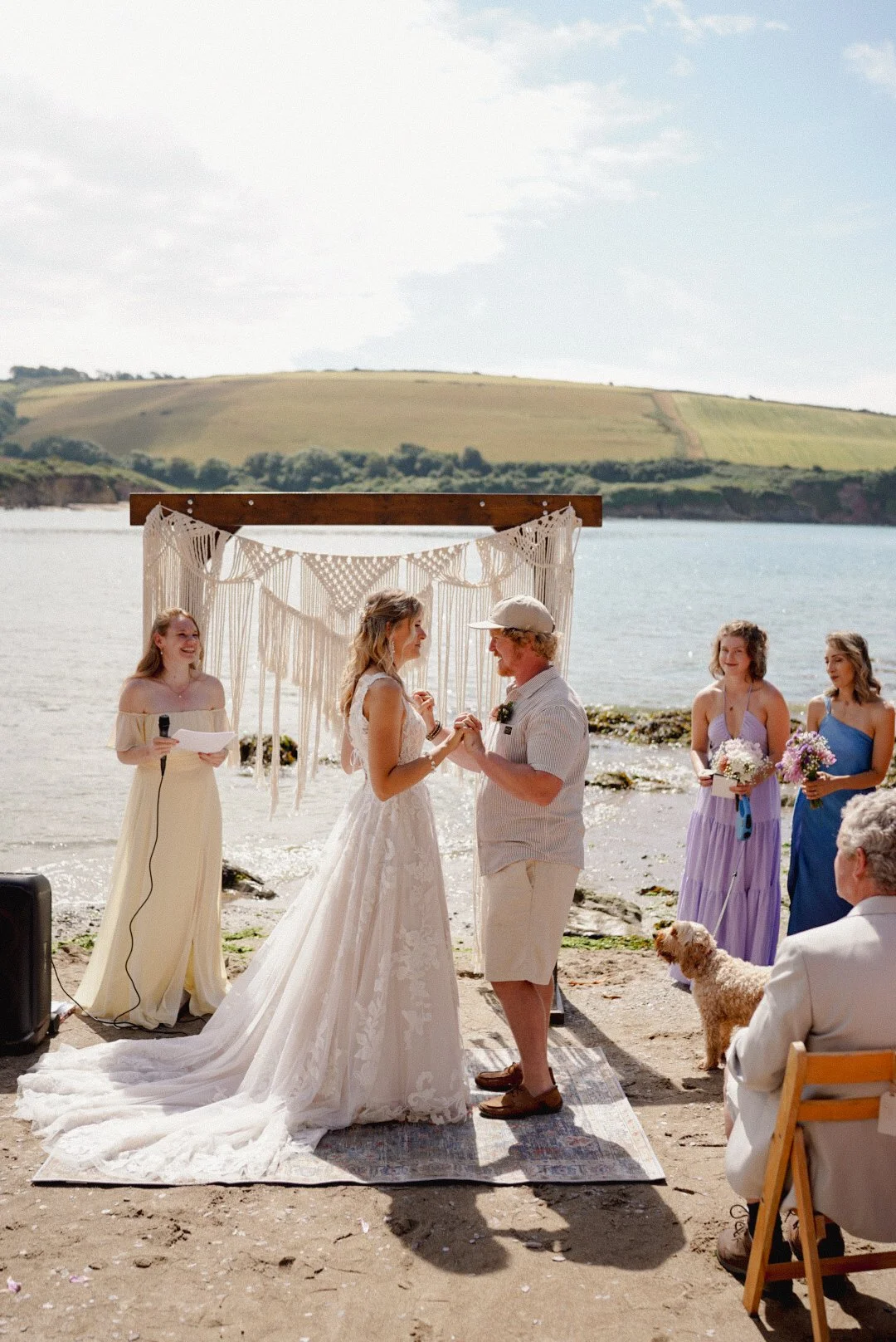 A couple getting married on a beach with friends and family, ocean, green hills, and cloudy sky in the background.