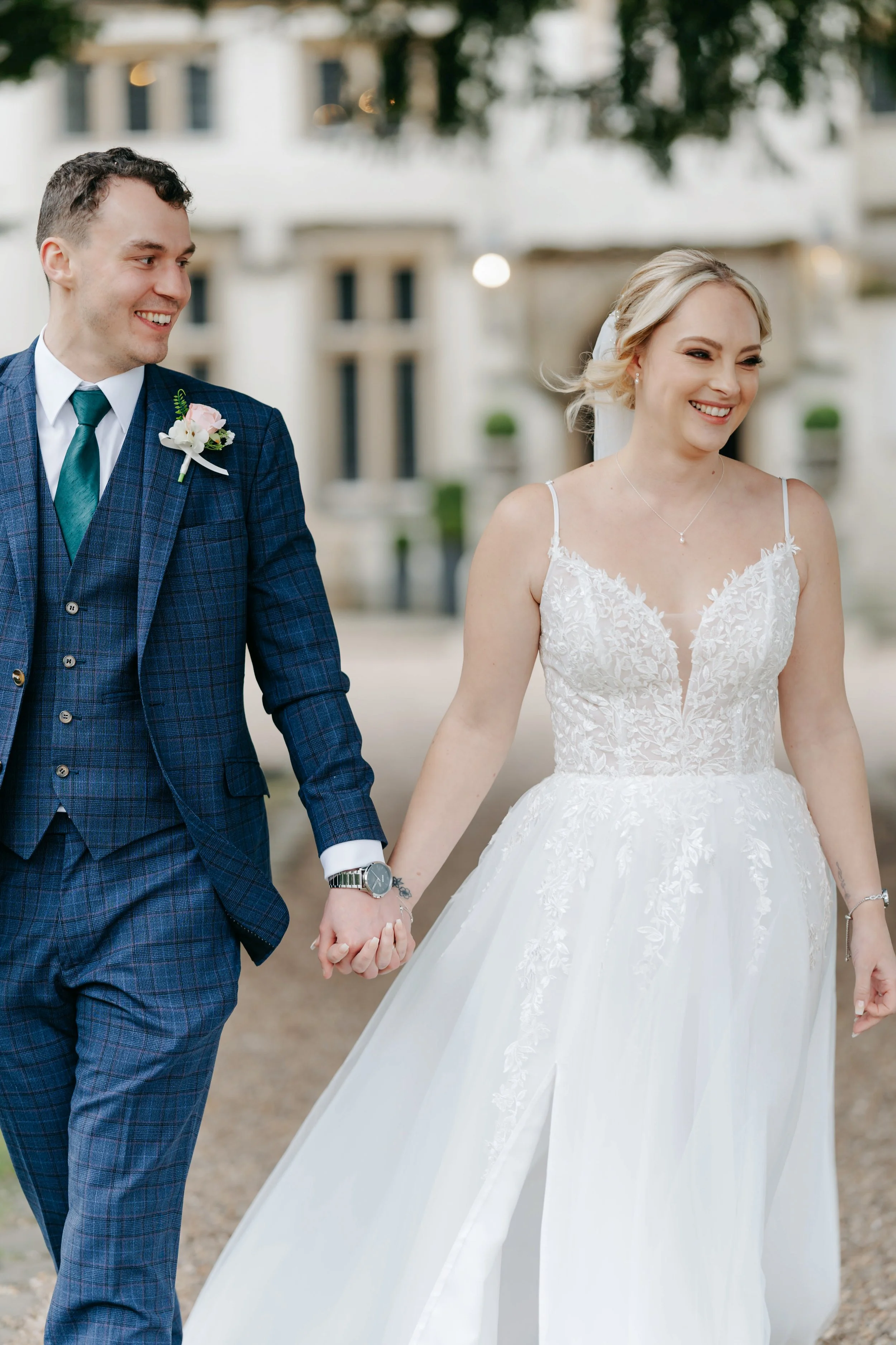 A bride and groom holding hands, walking outdoors during their wedding, smiling, with a blurred building background.