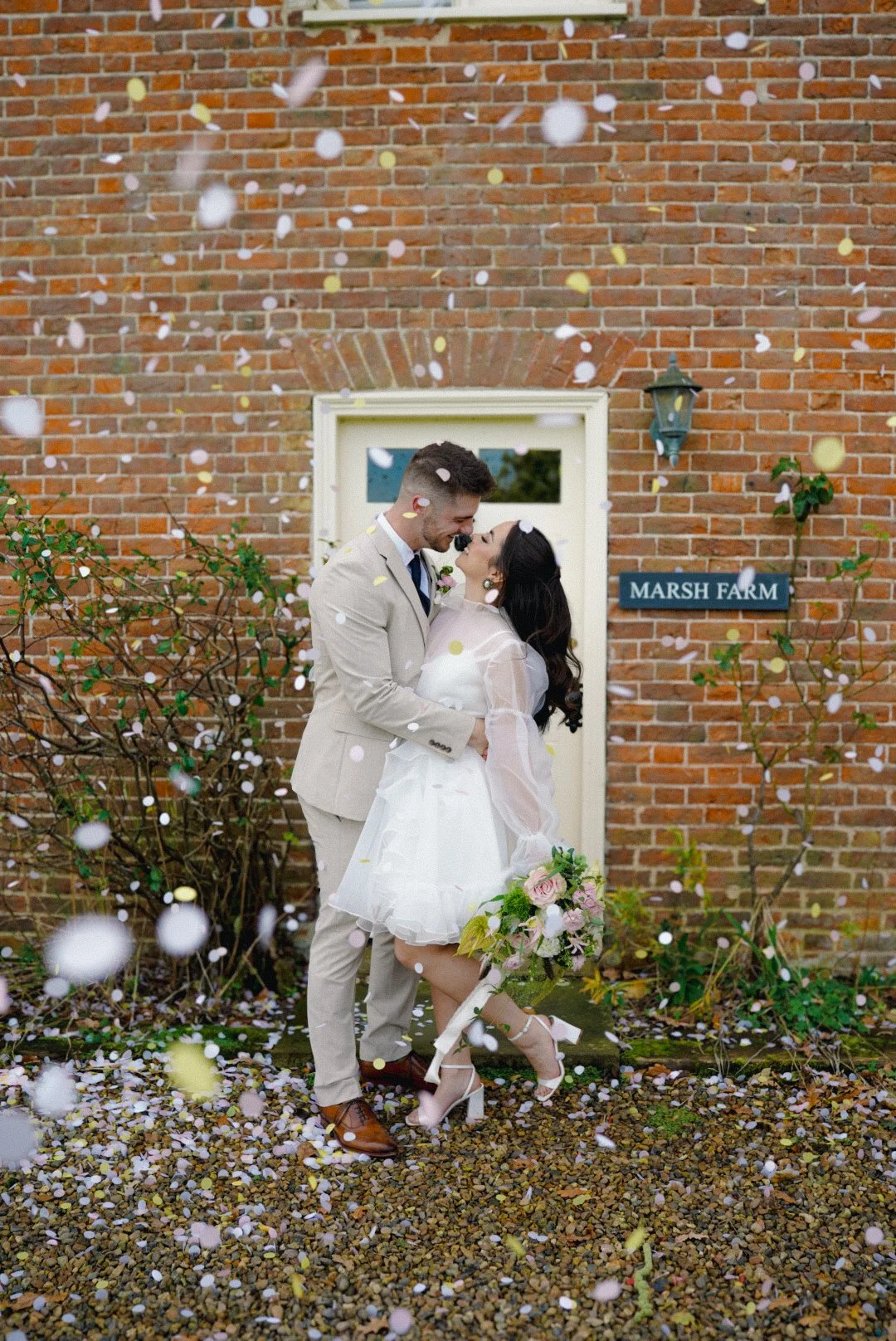 A newlywed couple is celebrating outside in front of a brick building, sharing a joyful moment while confetti falls around them. The bride is holding a bouquet of flowers and wearing a white dress, and the groom is in a light beige suit.