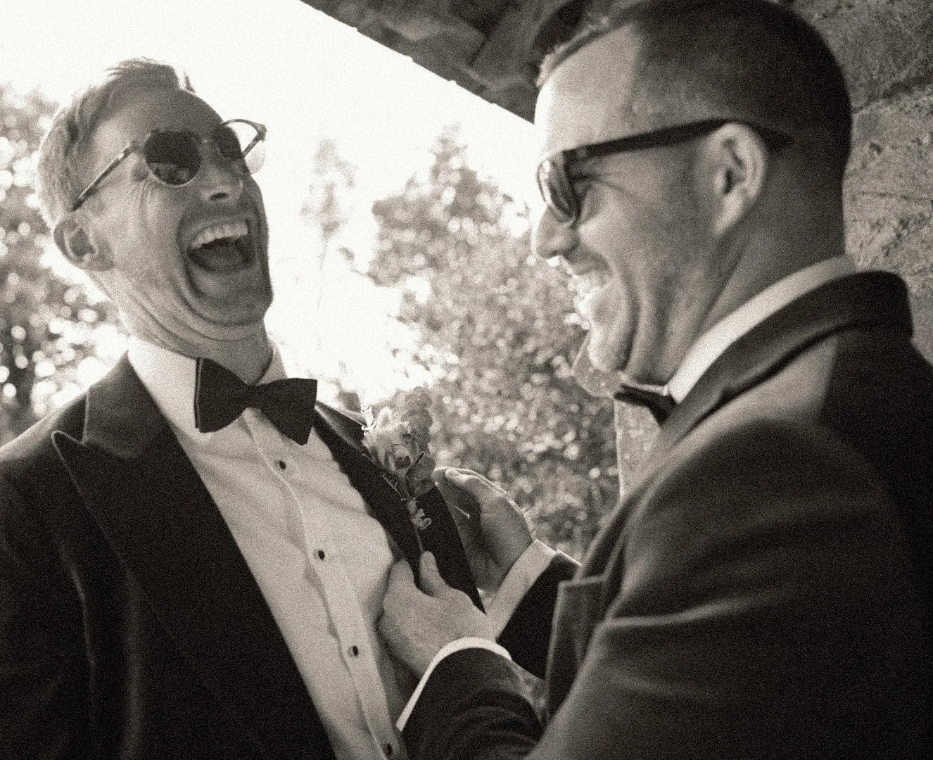 Two men in tuxedos, wearing glasses and bow ties, sharing a joyful moment outdoors, with one man holding a flower and the other holding his lapel.