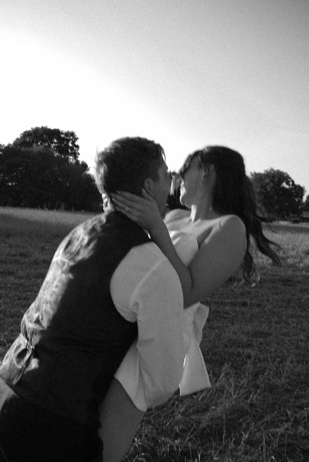 A black and white photo of a couple kissing outdoors in a grassy field during sunset, with trees in the background.