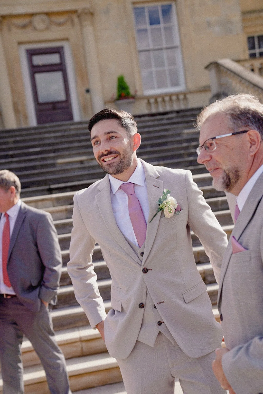 A groom and two other men in suits standing on the steps of a historic building with ornate architecture, smiling and engaging in conversation outside during the daytime.
