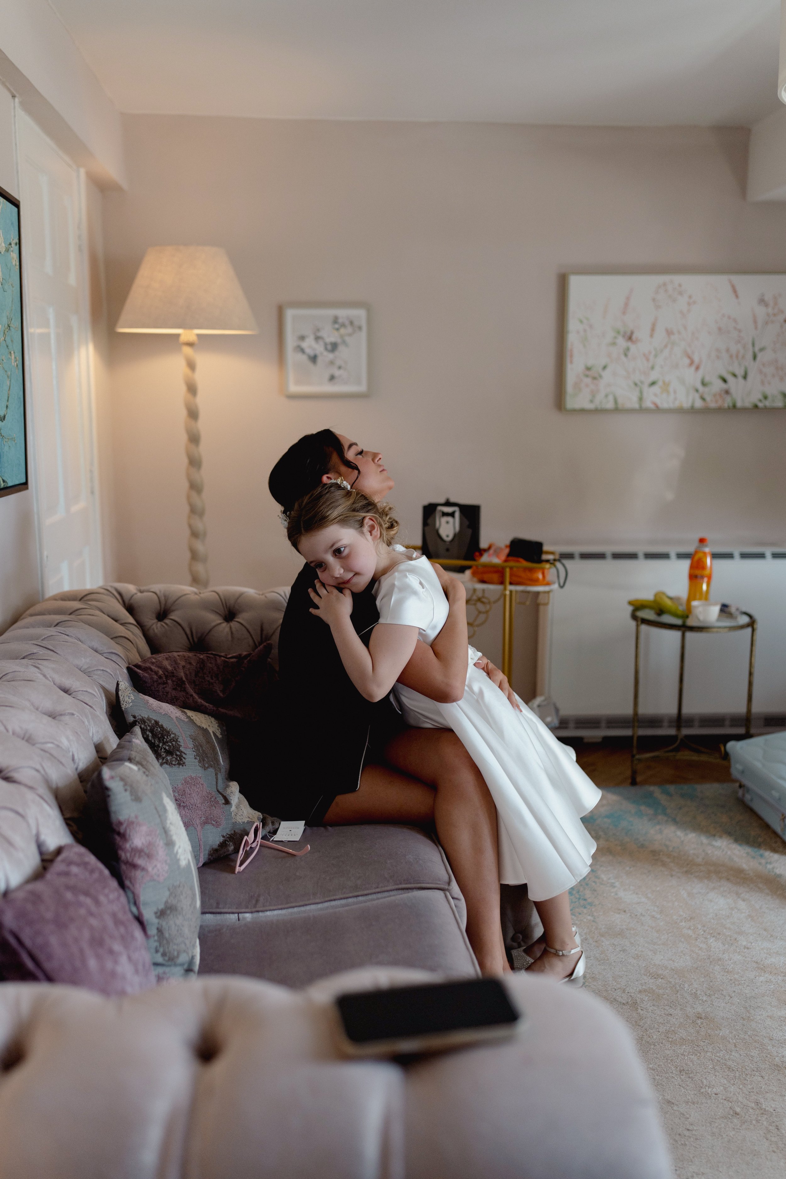 A woman and a young girl are sitting on a beige tufted sofa in a living room. The woman is hugging the girl who is resting her head on the woman's shoulder. The girl is dressed in a white dress, and the woman is dressed in dark clothing. The living r