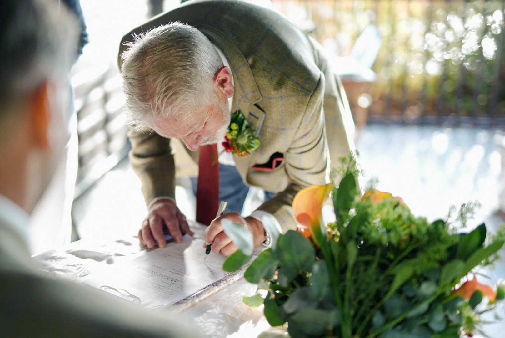An elderly man signing a document at a table with a floral arrangement, with a person blurred in the foreground and tree sunlight in the background.