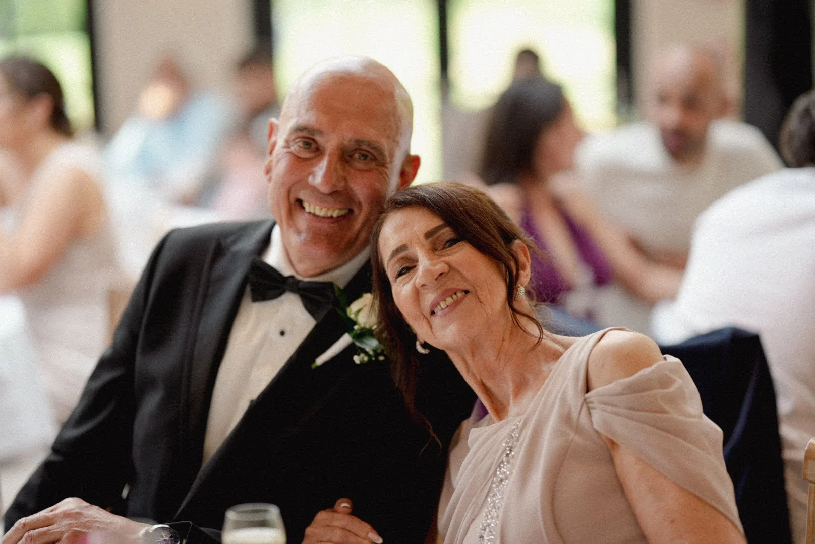 Elderly couple smiling, dressed in formal attire, at a wedding reception with guests in the background.