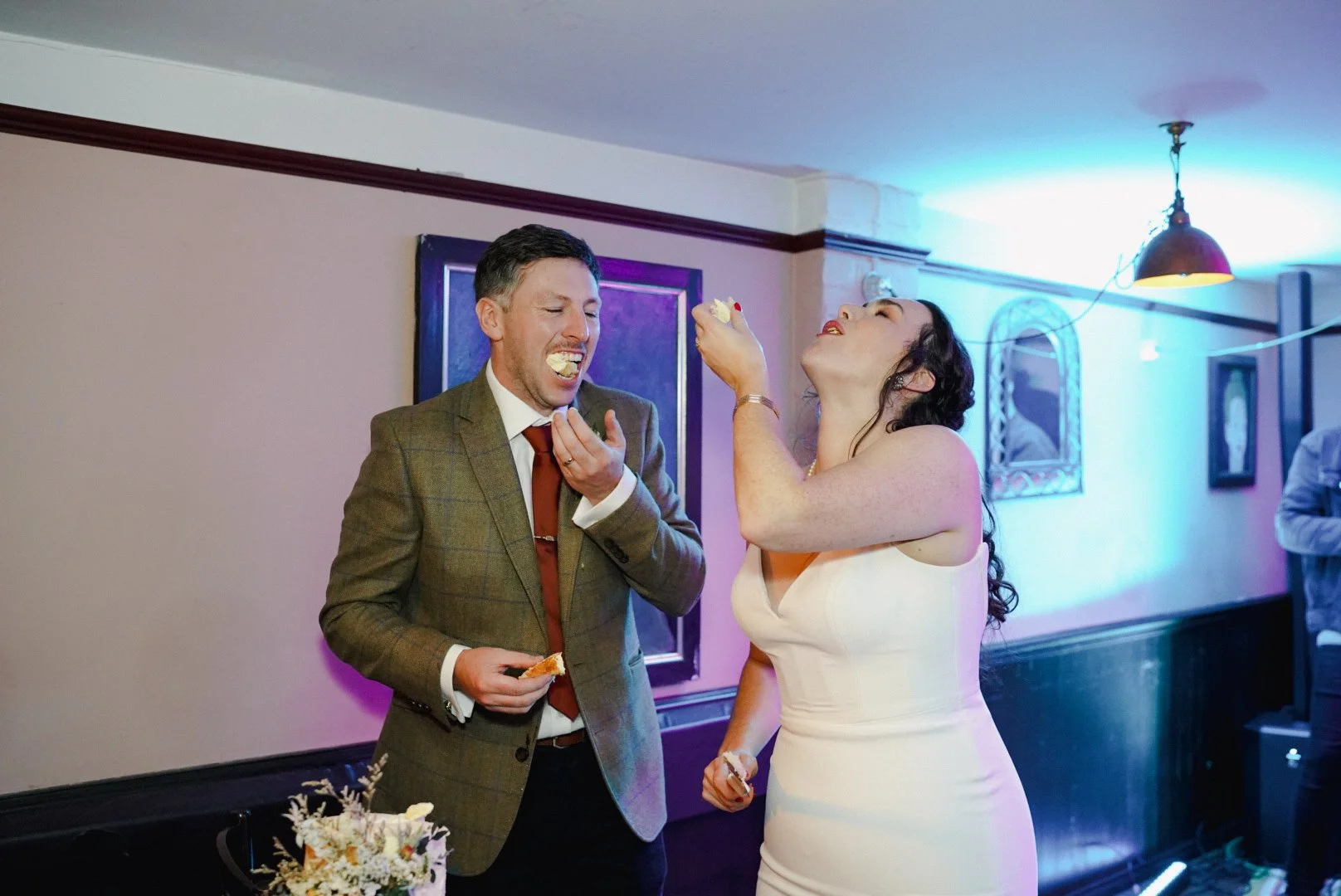 A man and woman at a celebration, sharing a slice of cake with cream filling, inside a decorated room with framed pictures on the wall.