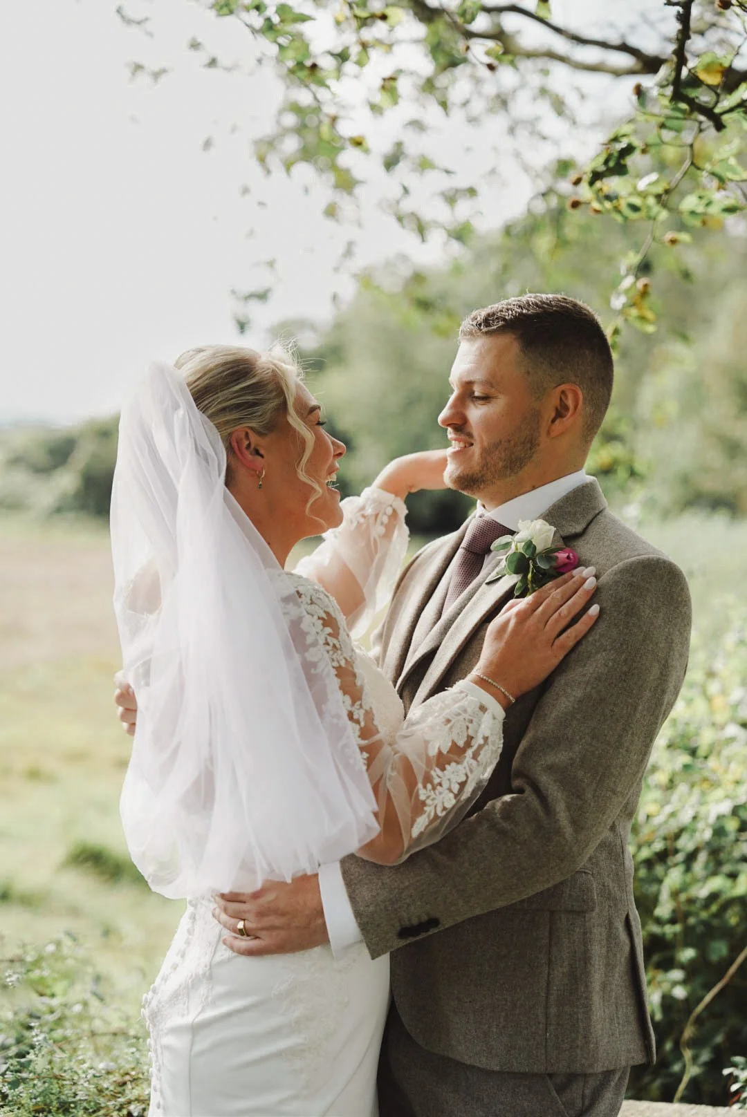 A bride and groom share a romantic moment outdoors during their wedding, smiling and looking into each other's eyes, with greenery and trees in the background.