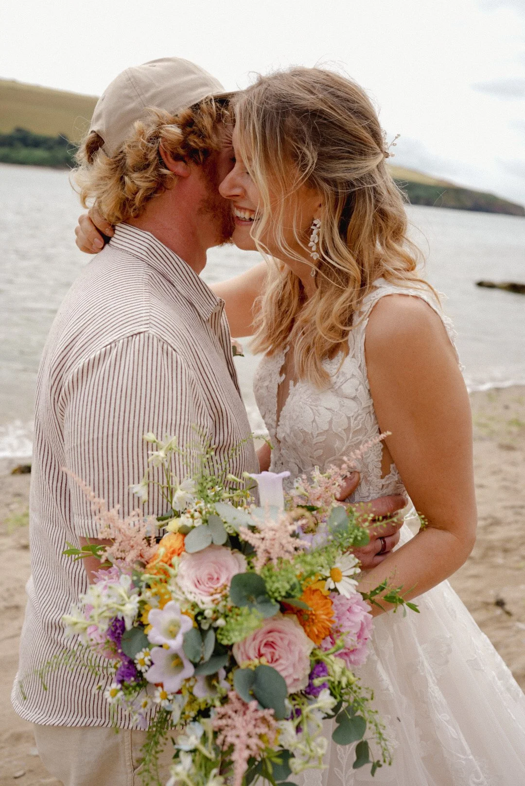 A couple in wedding attire embracing by the beach, with the bride holding a large colorful bouquet and smiling, while the groom leans in close.