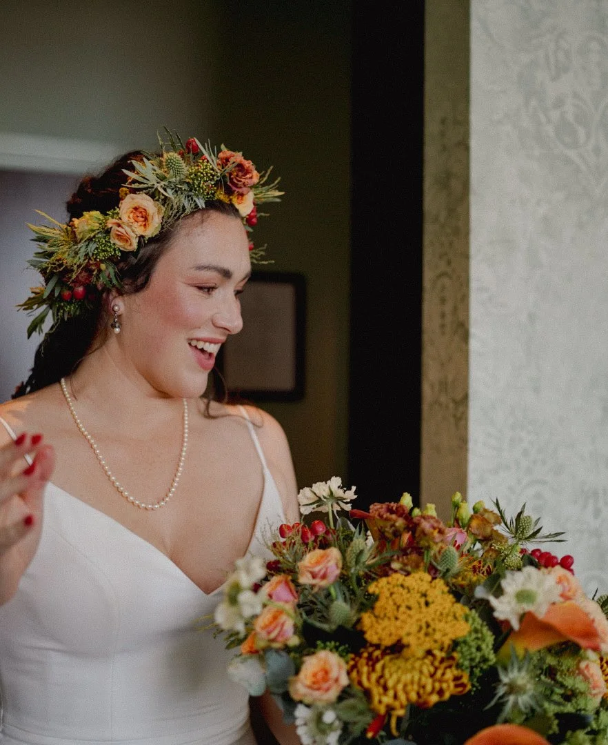 A woman with dark hair, wearing a white dress, pearl necklace, and floral crown, holding a bouquet of colorful flowers, smiling indoors.