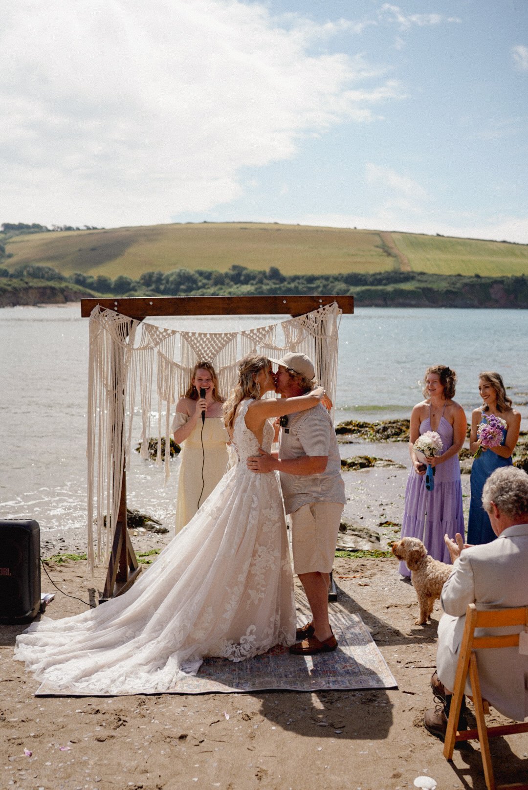 A bride and groom kissing during their beach wedding ceremony with bridesmaids and guests around them.