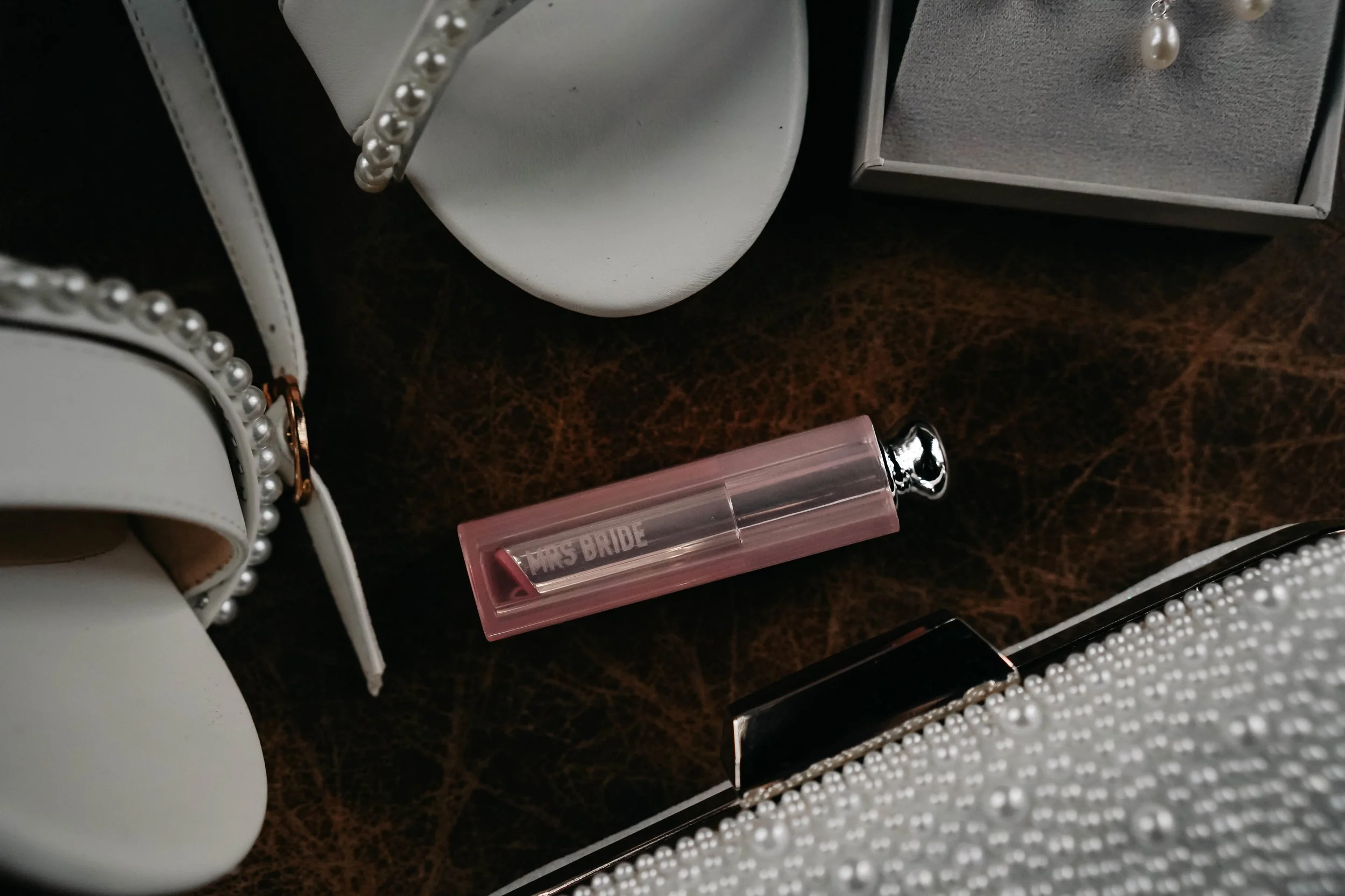 Close-up of accessories and items on a dark textured surface, including a white purse with pearl accents, a pink lipstick, a small gray jewelry box, and a gray round object.