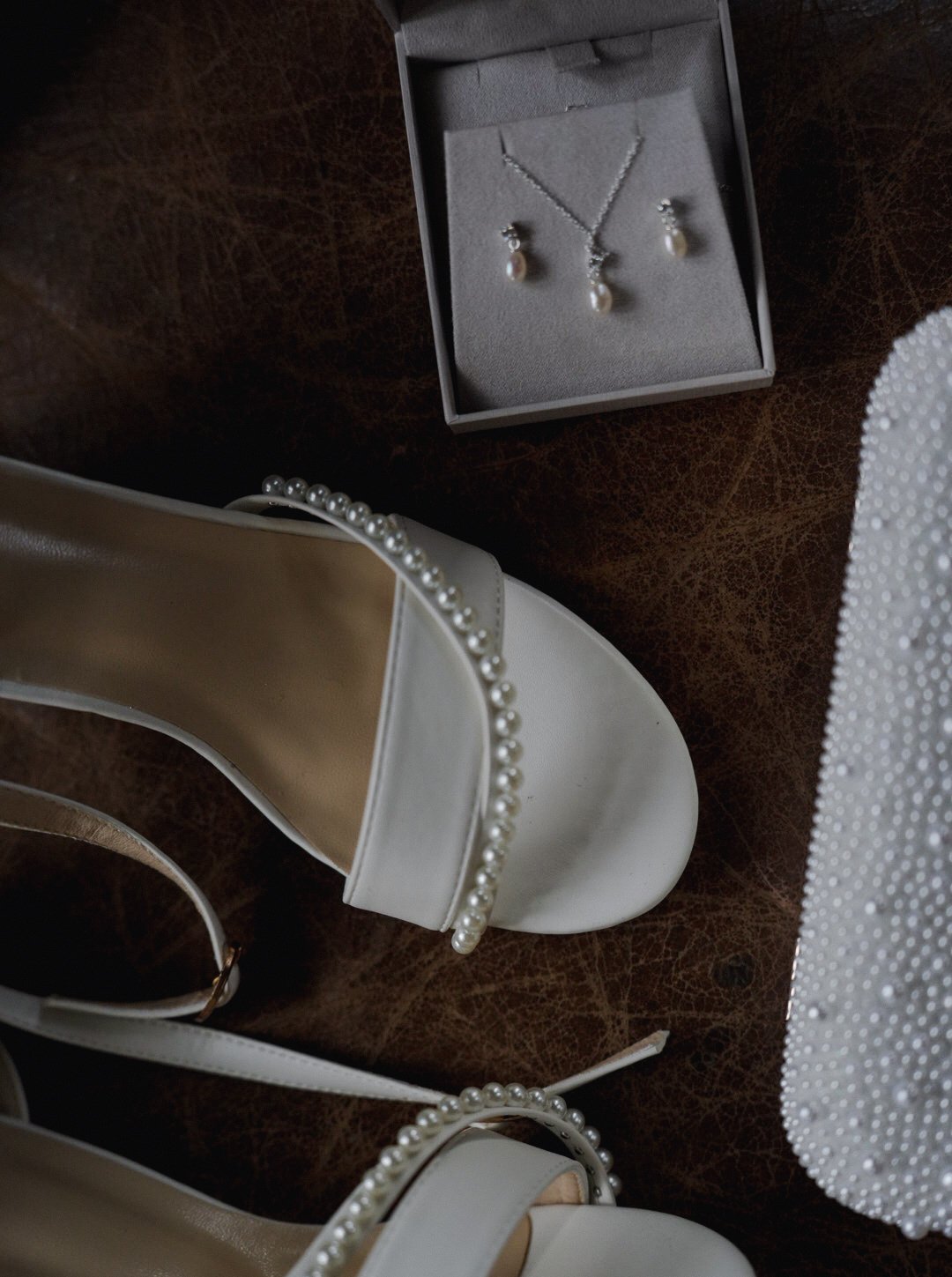 A pair of white wedding shoes with pearl embellishments, a pearl necklace and earrings in a jewelry box, and a white fabric with pearl beads, all arranged on a brown surface.