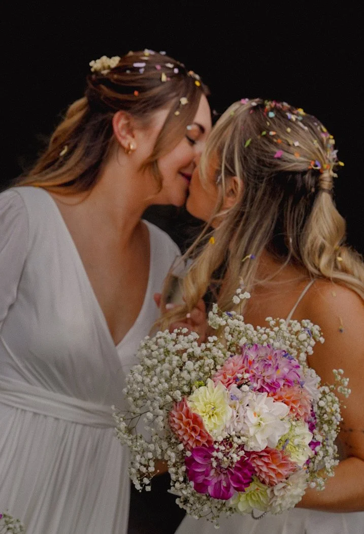 Two women with long, curly hair and confetti in their hair, sharing a kiss at a wedding. One woman is holding a colorful bouquet of flowers, including pink, white, and yellow blossoms, with baby's breath. Both women wear white dresses and the backgro