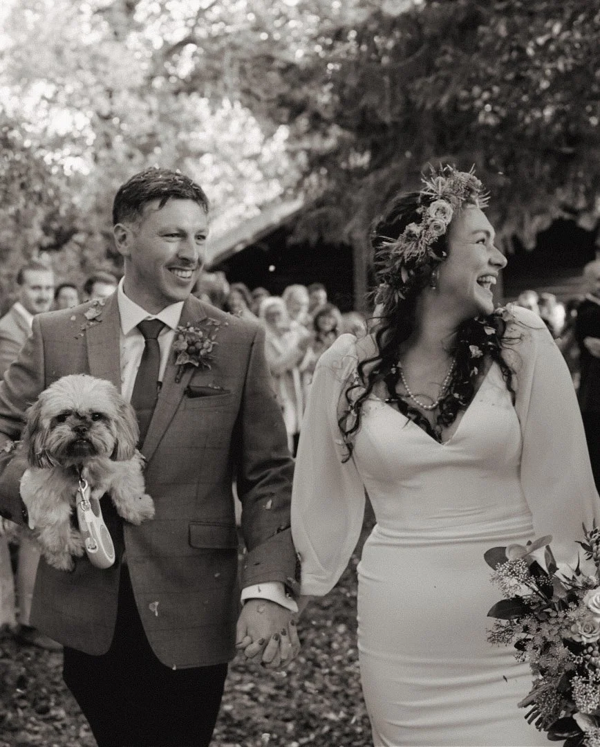 Black and white photo of a bride and groom holding hands during their outdoor wedding ceremony. The groom is wearing a suit and carrying a small dog. The bride is wearing a white dress and a floral crown, holding a bouquet, and is smiling.