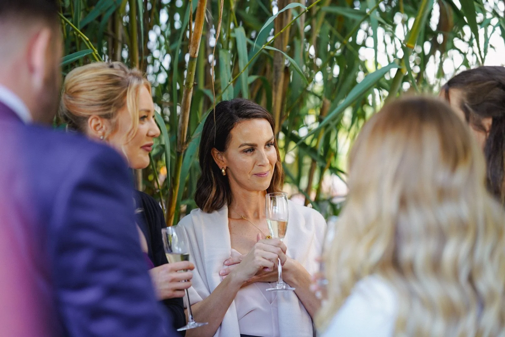 Group of people having a conversation, with woman holding a glass of champagne, in a lush green outdoor setting.