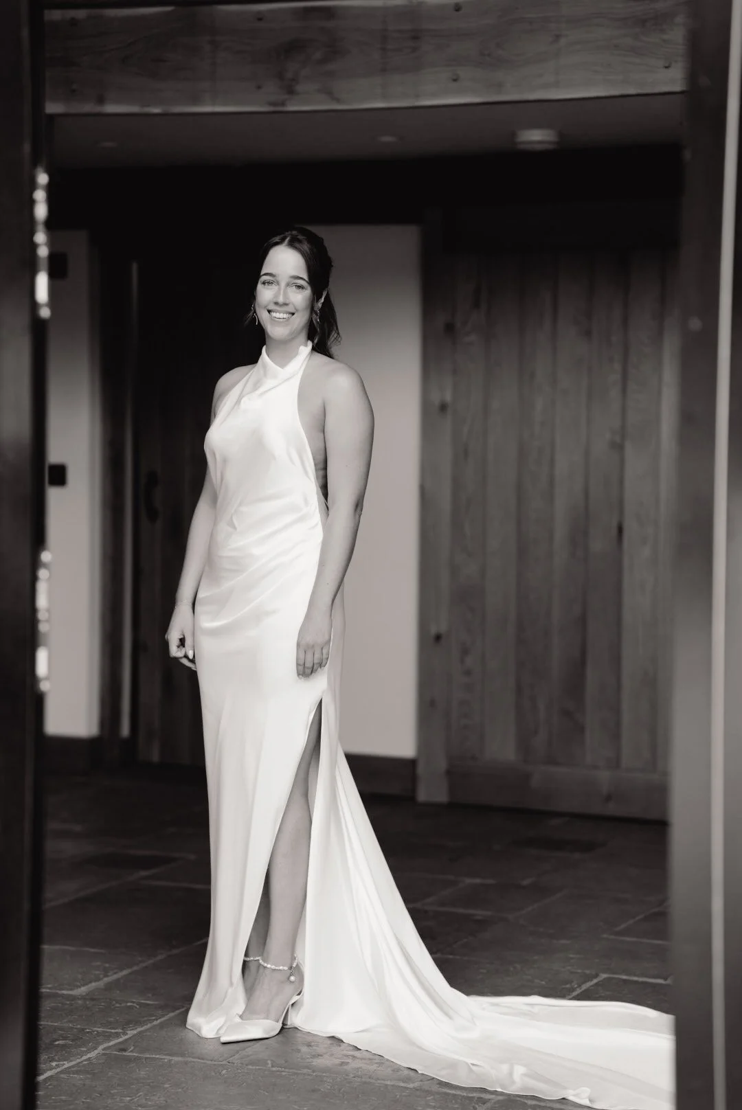 A woman in a white wedding gown standing in front of a mirror, smiling. The dress has a high slit and a train, with a sleeveless design, and she is wearing a pearl ankle bracelet and earrings.
