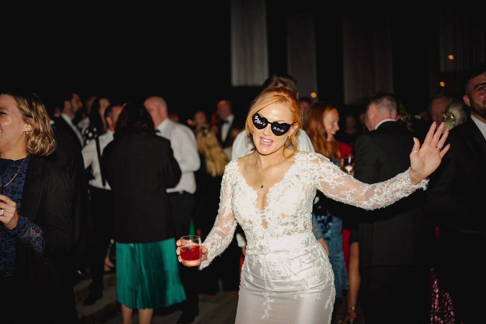 A woman with blonde hair wearing sunglasses and a white lace wedding dress dancing at a wedding reception, holding a glass of red drink.