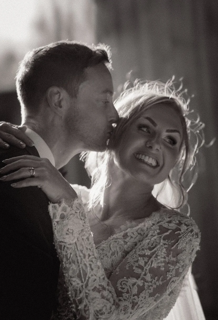 A black and white photo of a bride and groom dancing at a wedding reception, with the groom kissing the bride on her cheek. The bride is smiling and wearing a lace wedding dress.