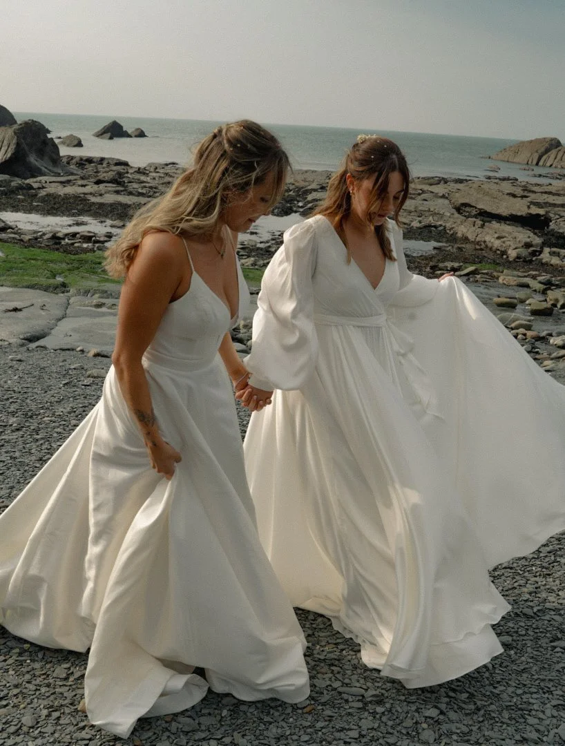 Two women in white wedding dresses holding hands and walking on a rocky beach with ocean in the background.