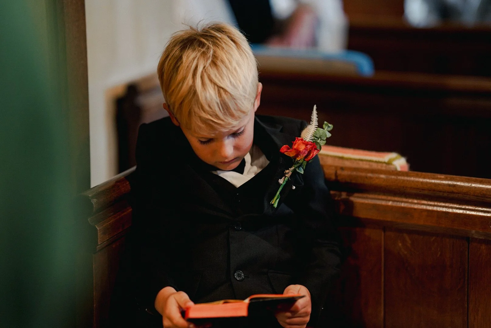 A young boy with blond hair sitting on a wooden pew, reading a book, wearing a black suit with a flower on his lapel.