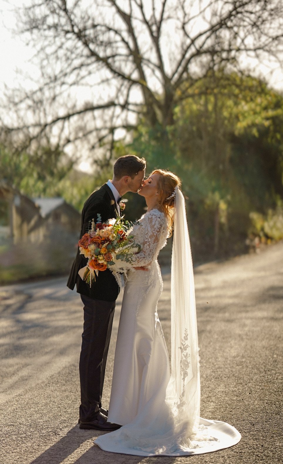 A bride and groom sharing a kiss outdoors during sunset, with the bride in a white lace wedding gown holding a colorful bouquet, and the groom in a black tuxedo, in front of a leafless tree.