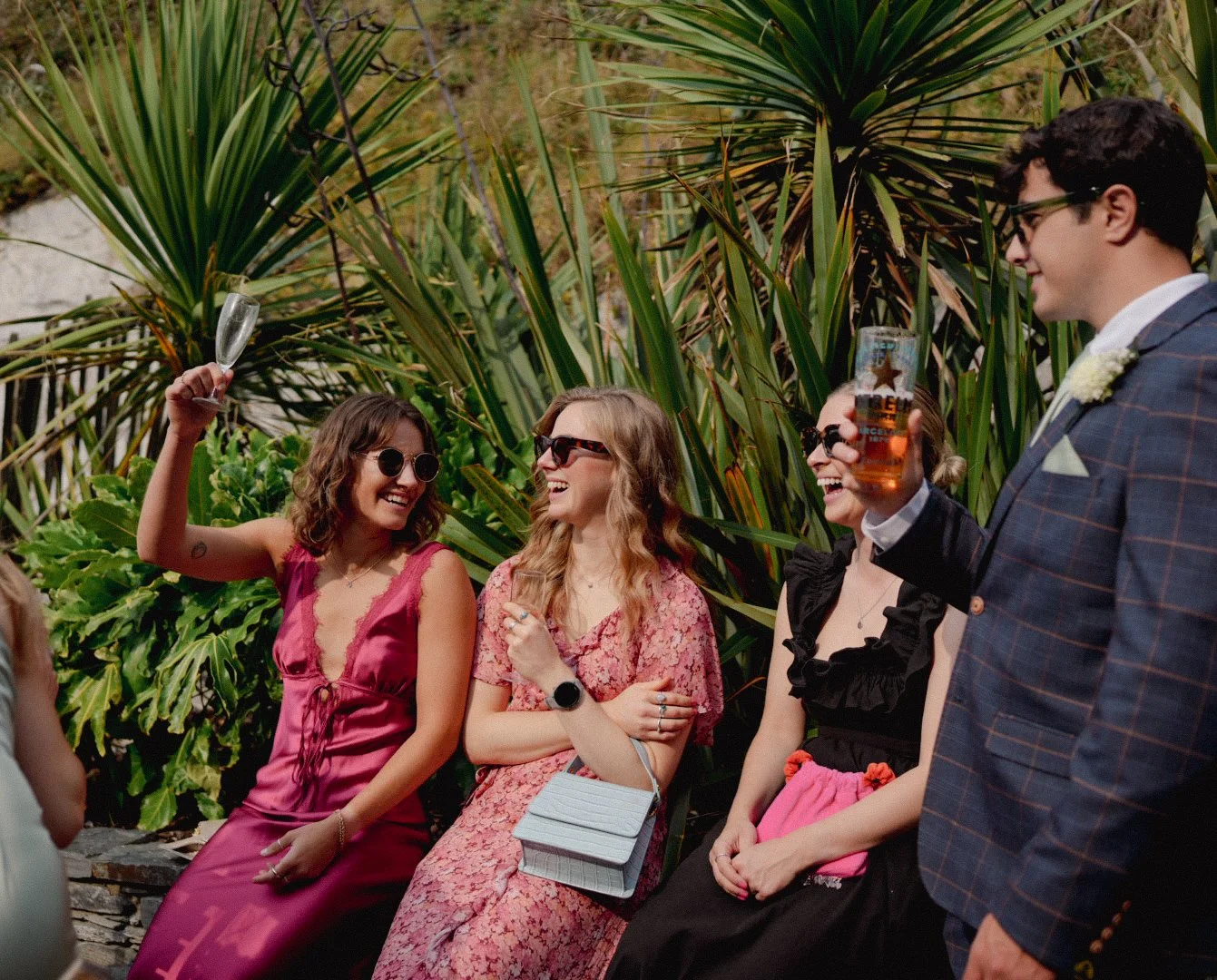 Four friends at an outdoor party celebrating, holding drinks, smiling, and talking, with tall green plants in the background.