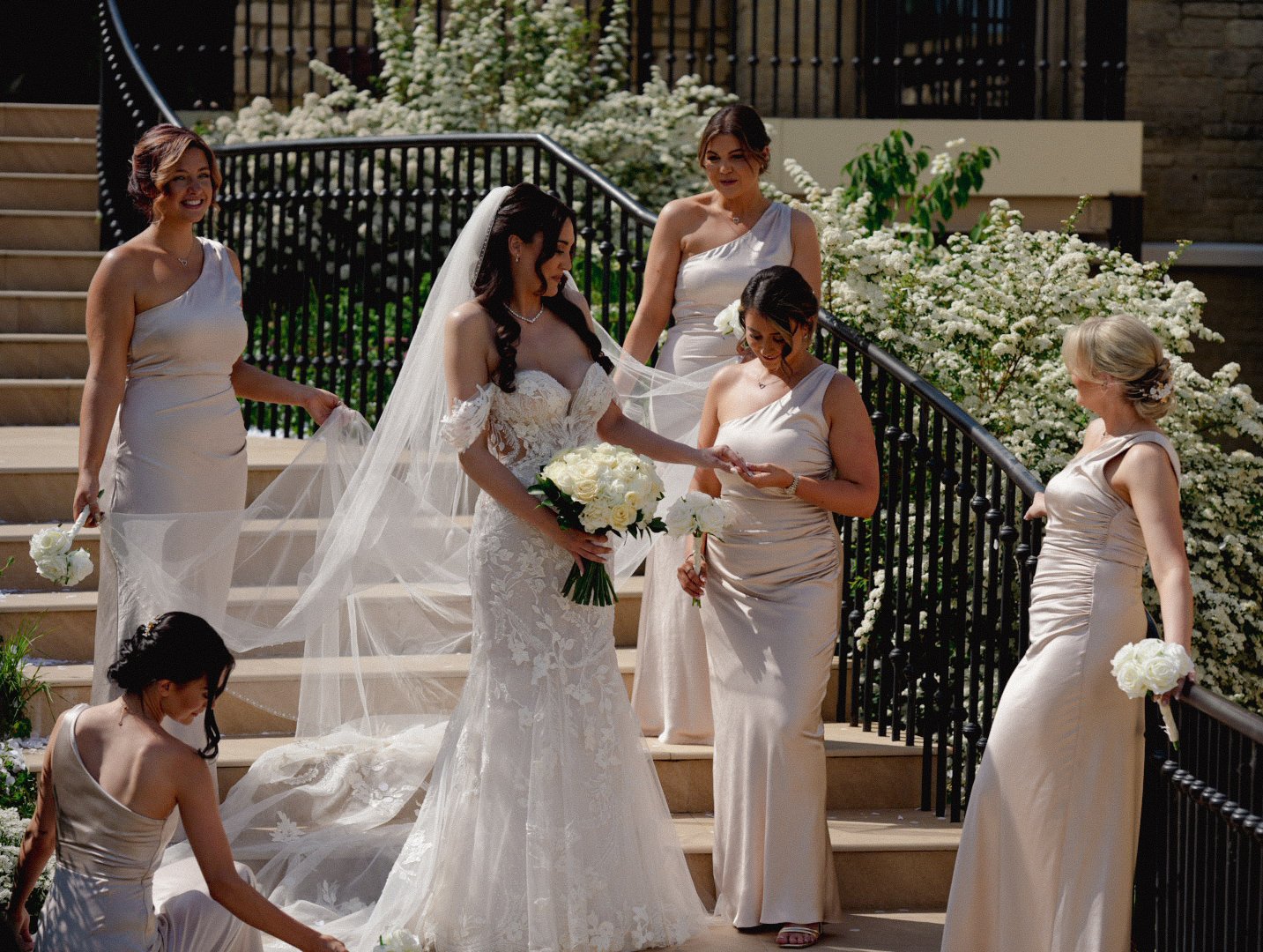 Bride in a white lace wedding gown with veil holding a bouquet, surrounded by bridesmaids in beige satin dresses and holding white flowers, on outdoor steps decorated with white flowers and greenery.
