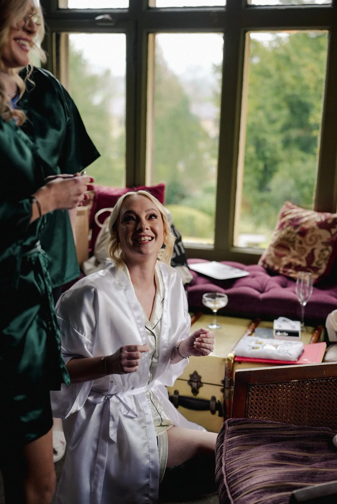 A woman in satin pajamas sitting on a bench, smiling and looking up, with another woman in a dark robe standing beside her, inside a cozy room with large windows and a view of greenery outside.