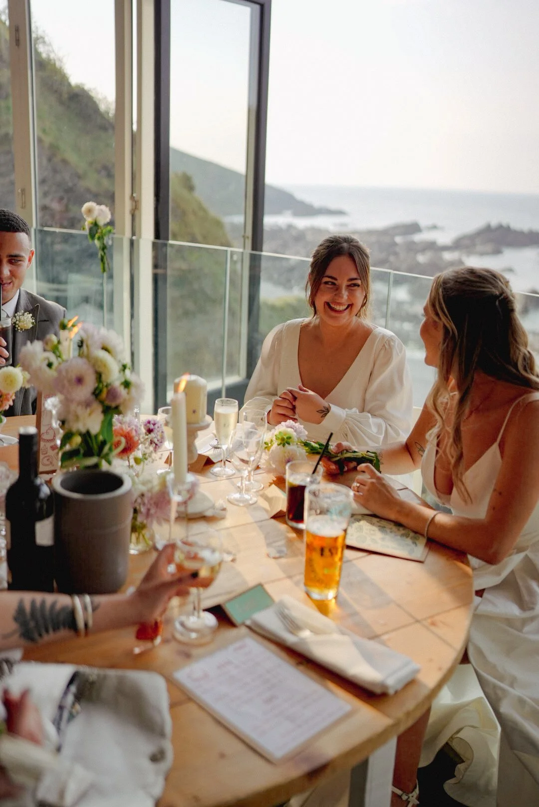 People celebrating at a wedding reception, sitting around a table with floral arrangements, candles, glasses of beverages, and a scenic ocean view through large windows in the background.