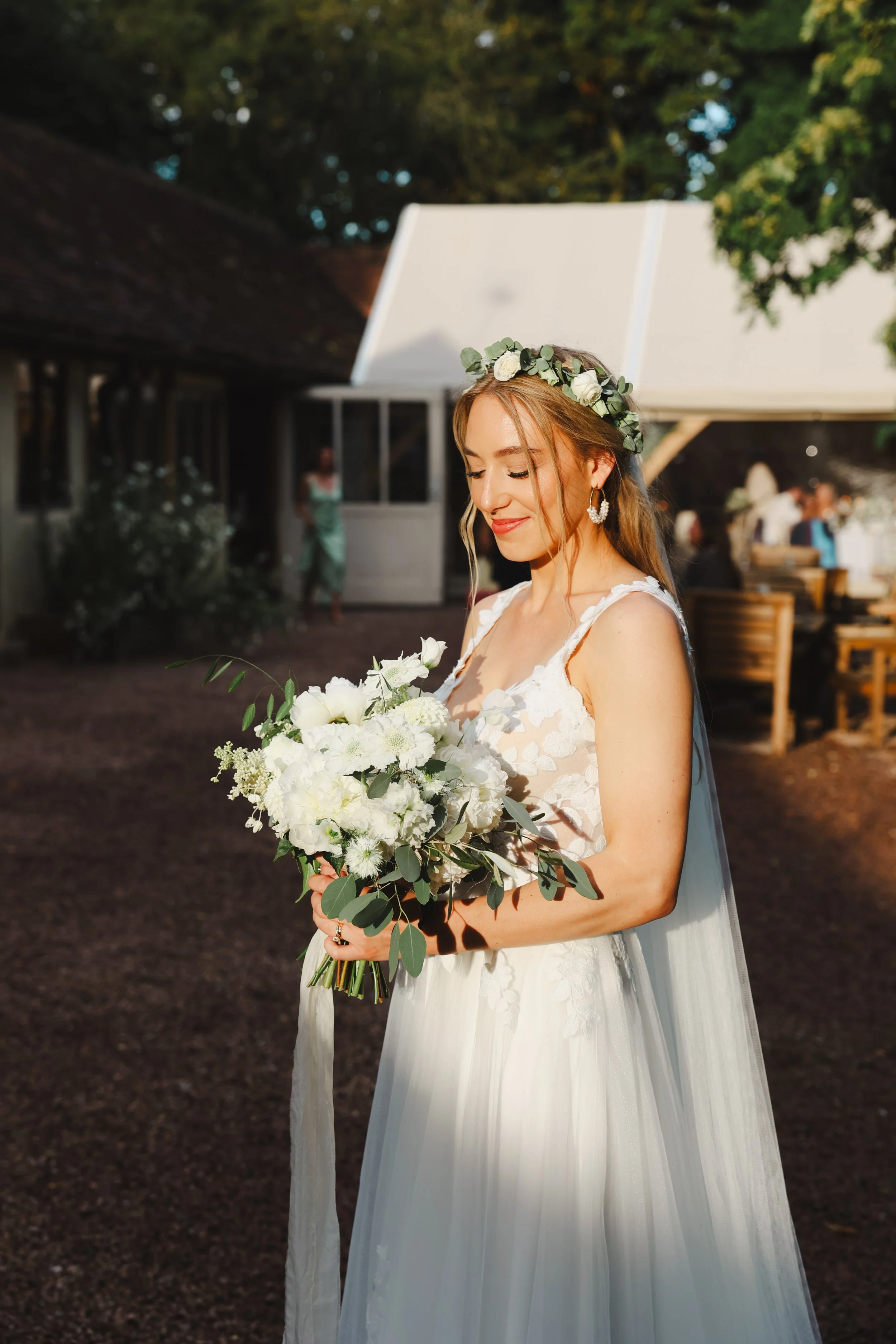 Bride in a white wedding dress holding a bouquet of white flowers and greenery, standing outdoors with a wedding venue and guests in the background.