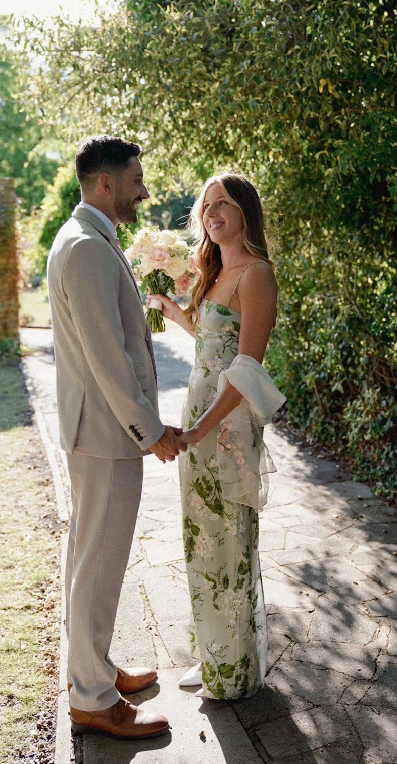 A couple holding hands outdoors, the woman giving a bouquet to the man, with greenery and trees in the background.