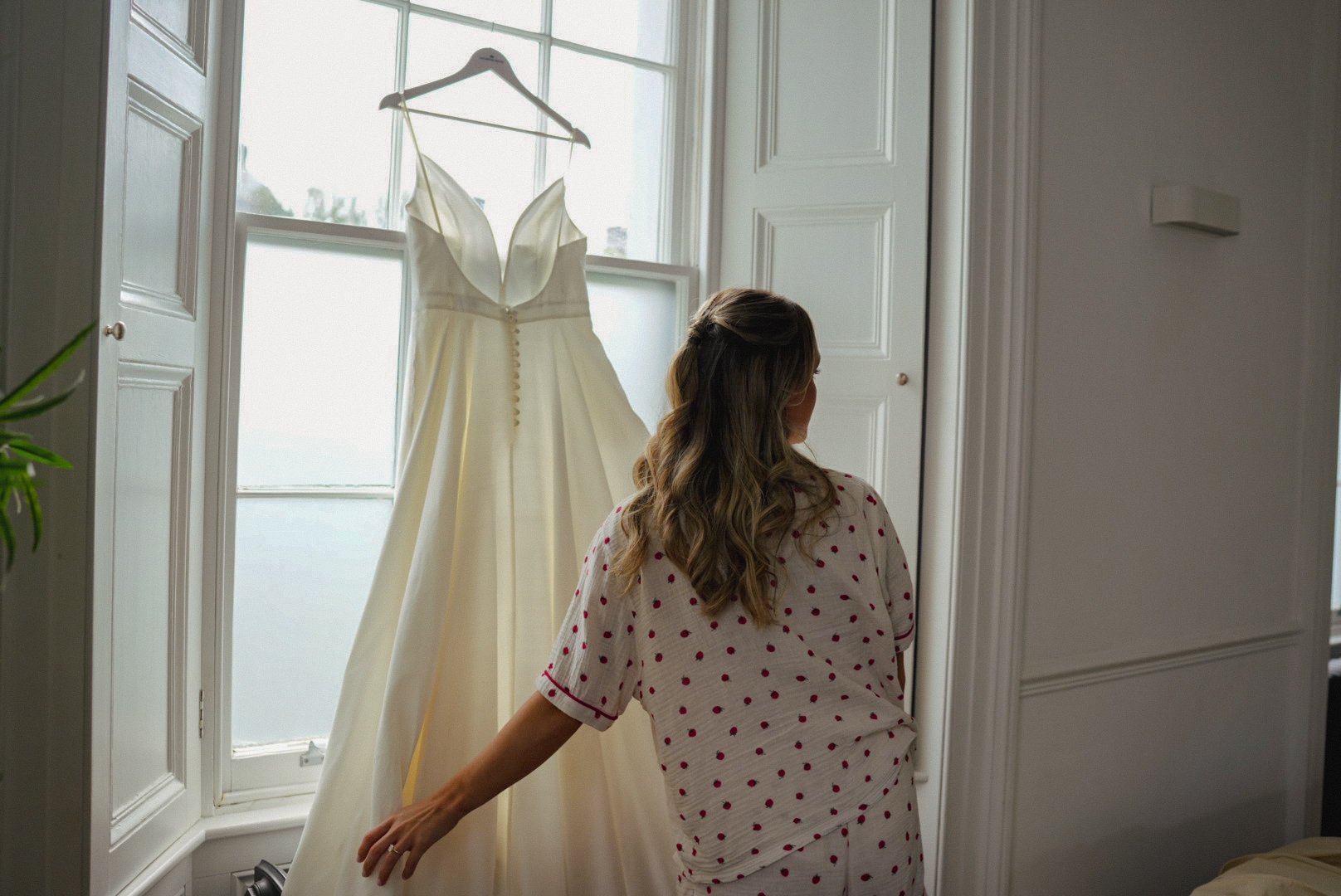 A woman in pajamas looking at a wedding dress hanging in front of a window.
