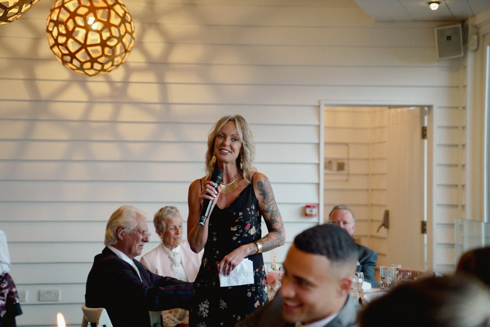 A woman with blonde hair, tattooed arms, and a floral dress holding a microphone, speaking at a social event in a room with white wood-paneled walls and a geometric chandelier.