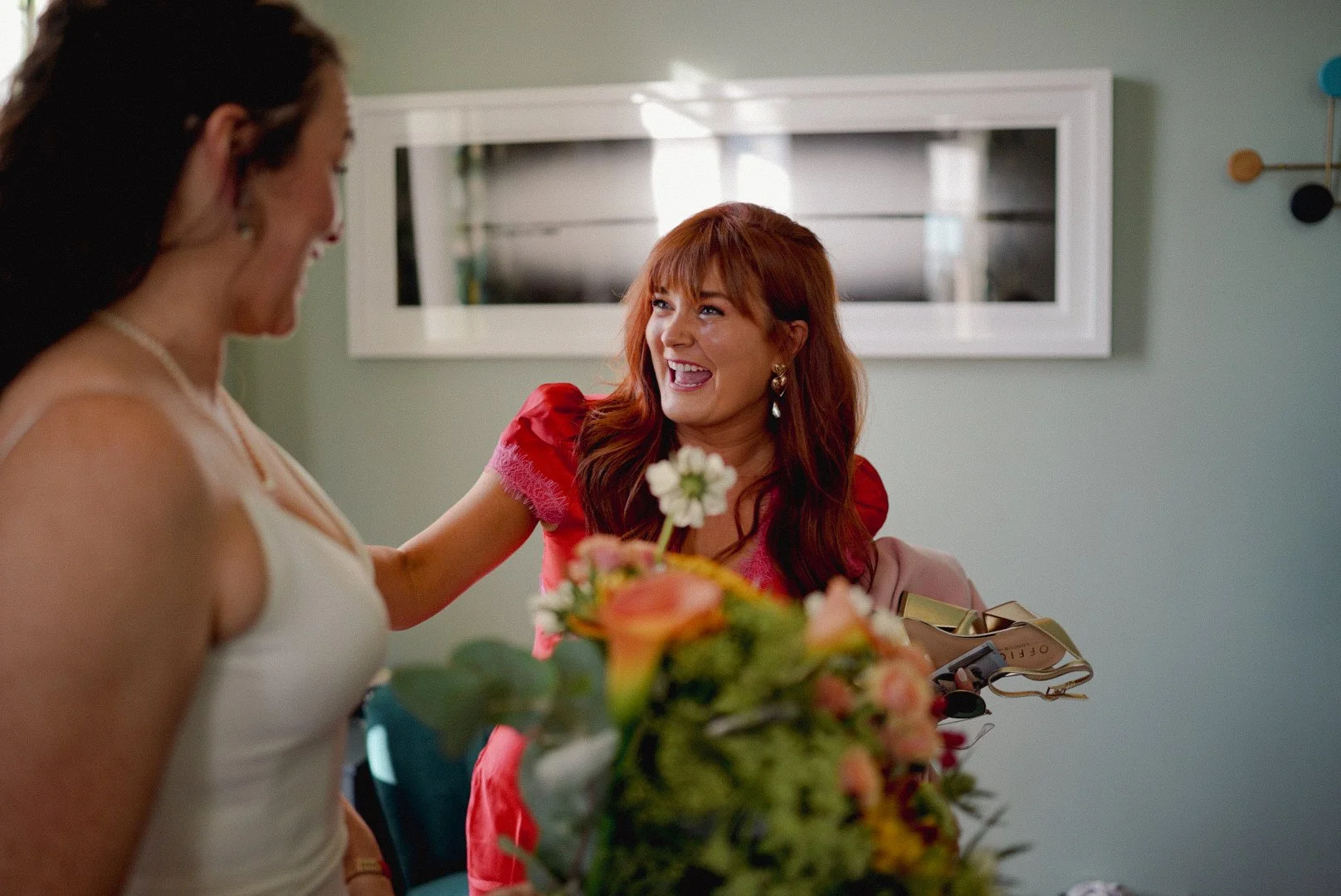 Two women smiling and talking, one with a bouquet of flowers in front of her, in a room with a mirror on the wall.