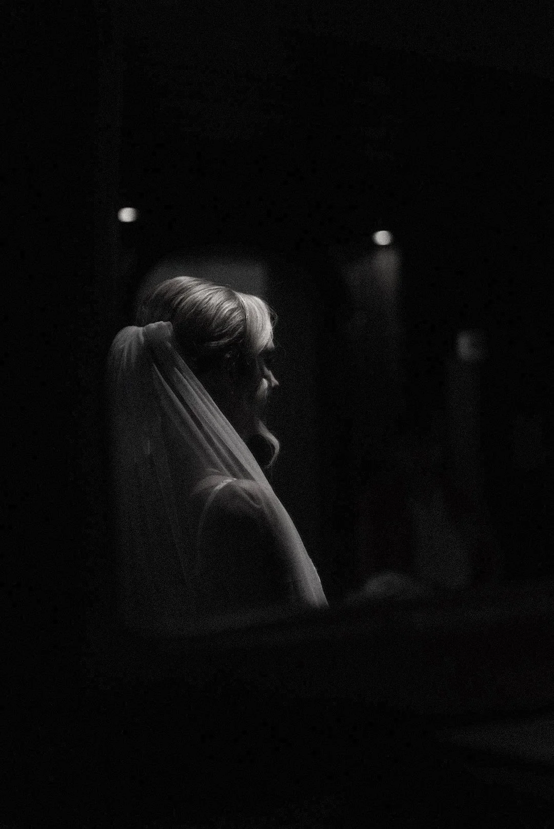 A black and white photo of a woman with a veil and hairstyle, sitting in a dark room with faint lights in the background.