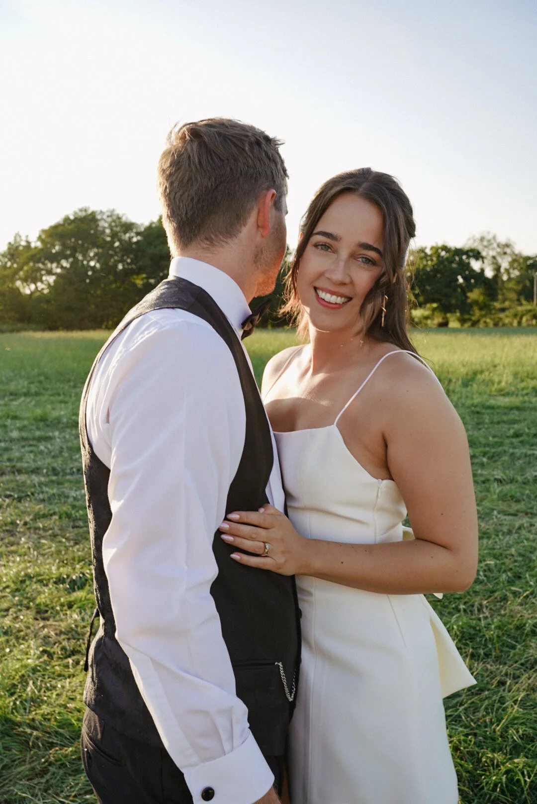 A smiling woman in a white dress standing in a grassy field with a man in a tuxedo, both during sunset.