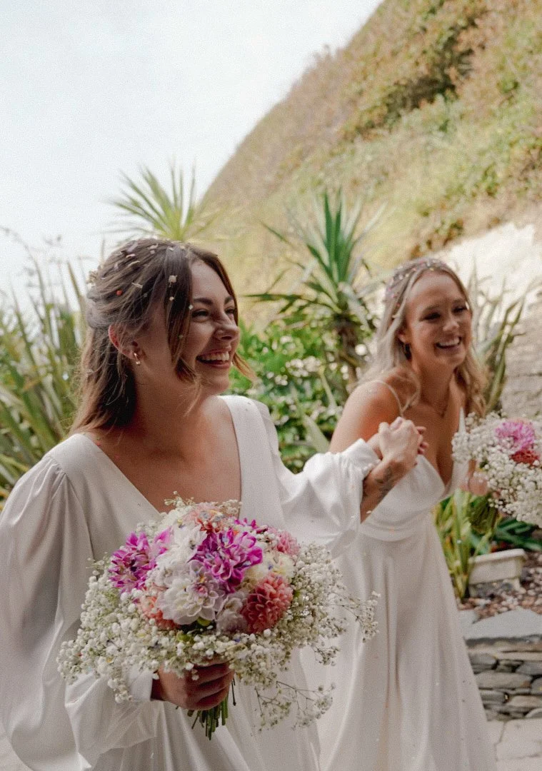 Two women in white wedding dresses holding bouquets of colorful flowers, smiling, in a lush outdoor setting.