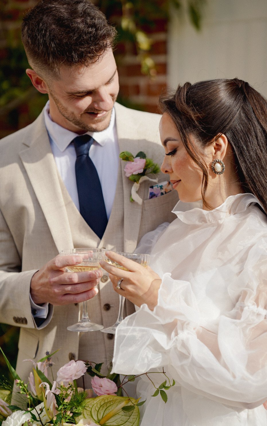 A newlywed couple sharing a toast at their wedding celebration, with the groom dressed in a beige suit and the bride in a white dress, holding champagne glasses, surrounded by flowers.