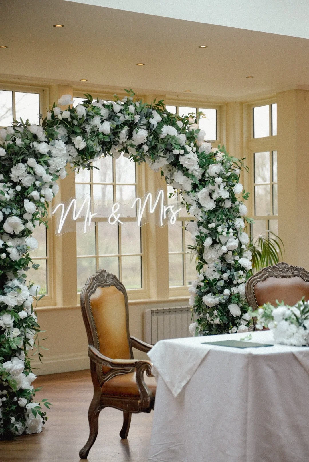 Wedding reception area with a floral arch that has a neon sign saying "Mr & Mrs" and vintage chairs around a table with a white tablecloth, set in a room with large windows.