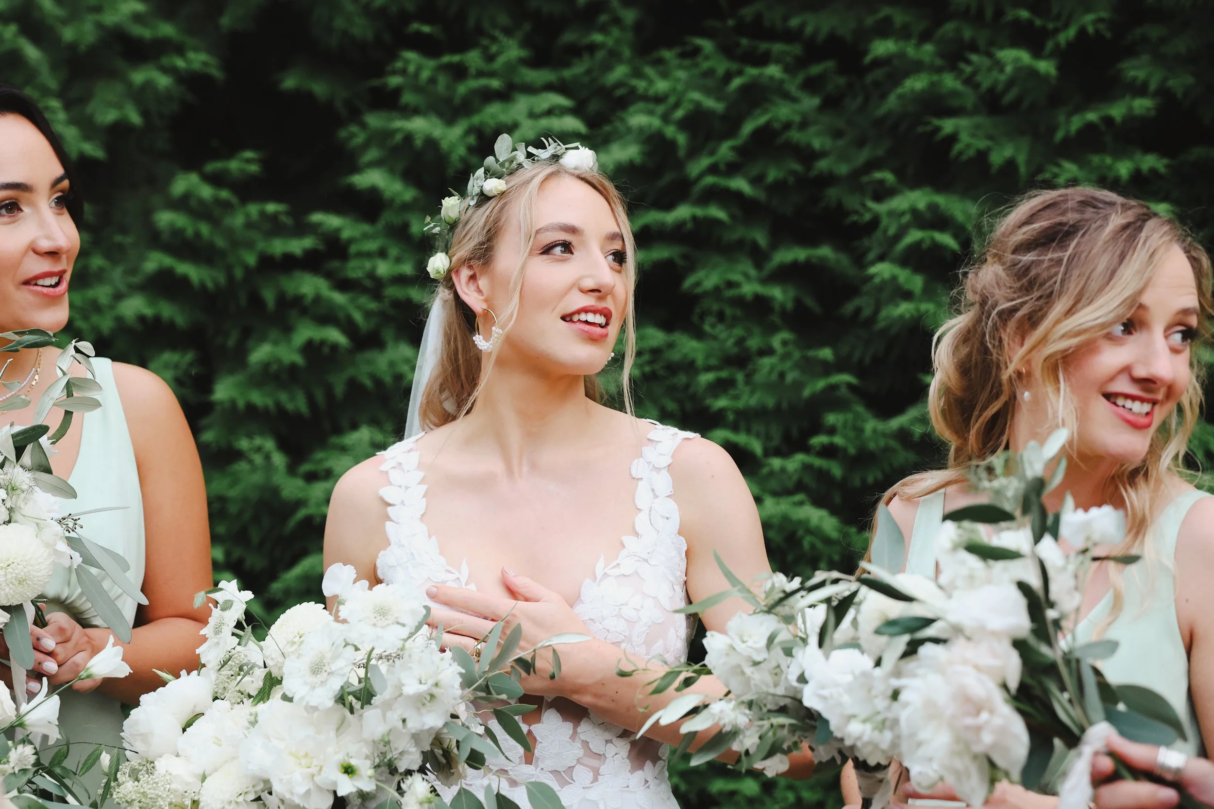 A group of women in bridal dresses holding bouquets of white flowers, standing outdoors with green foliage in the background.